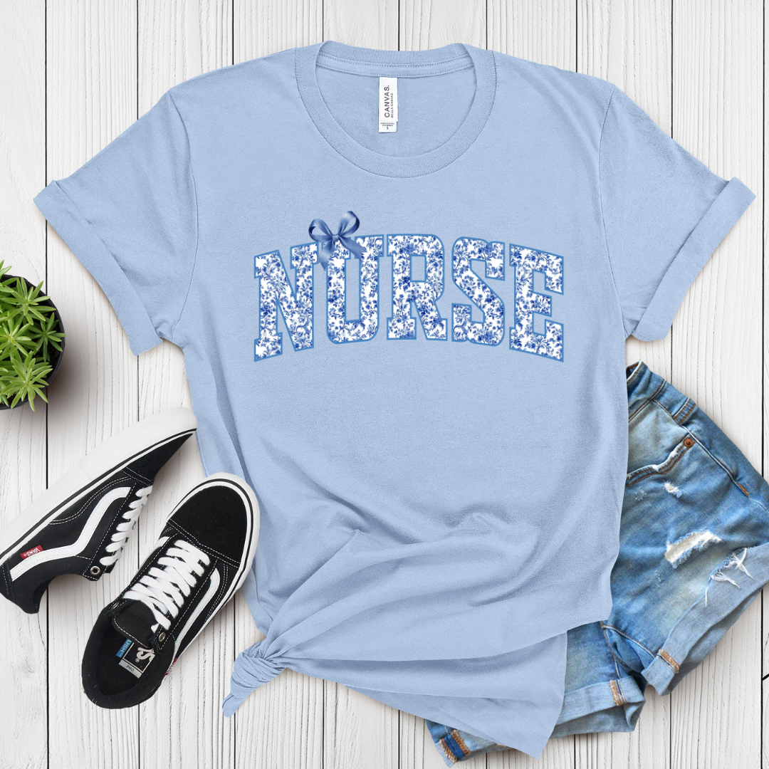 Light blue t-shirt with 'NURSE' printed on it, paired with denim shorts and sneakers on a wooden surface.