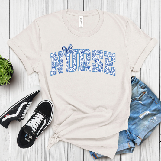 A white crew neck t-shirt with the word 'NURSE' printed in blue varsity-style text, adorned with blue bows, laid flat with a pair of blue jeans, a pair of black and white sneakers, and a green plant in the background.
