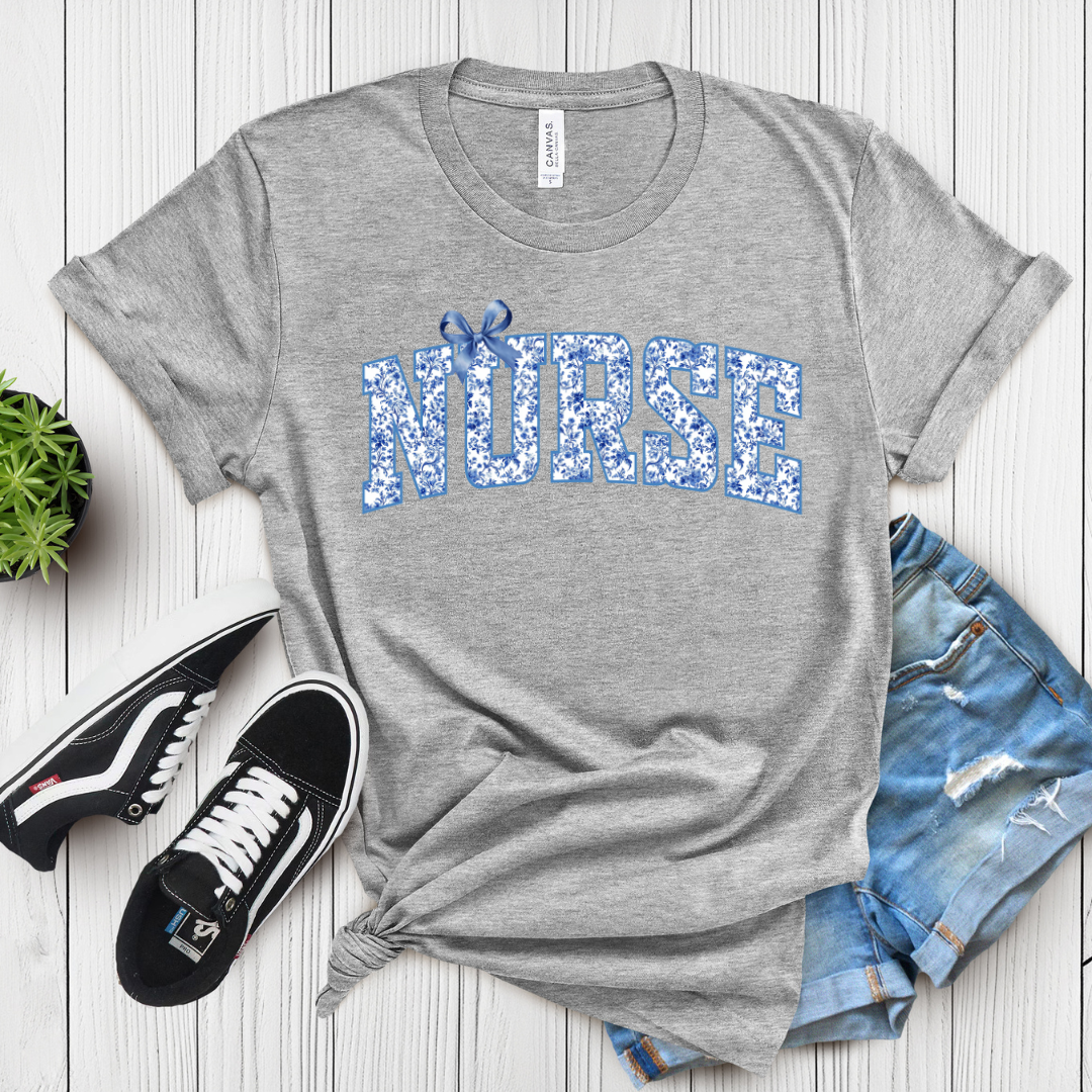 Gray t-shirt with 'NURSE' printed in blue floral letters, paired with black sneakers and denim shorts on a wooden surface.