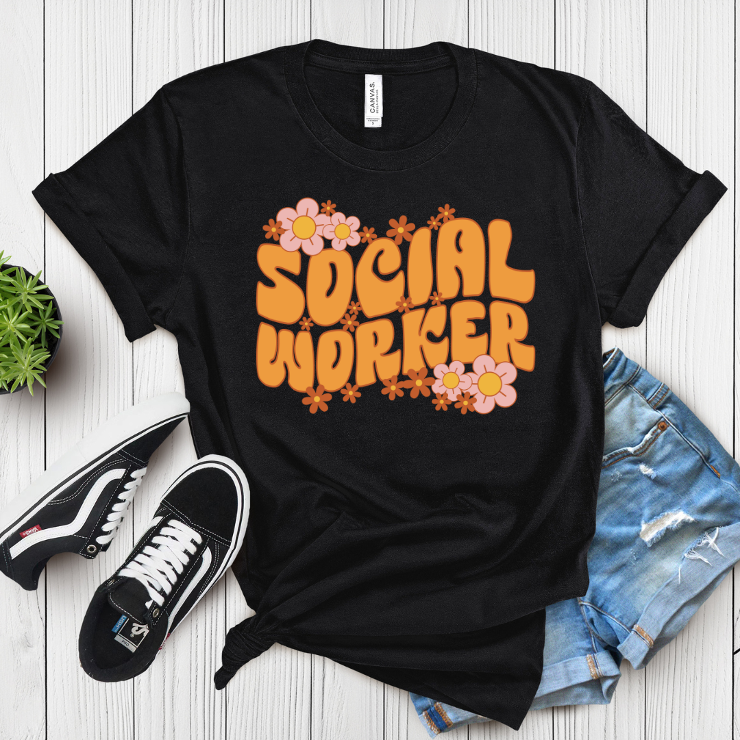 Black t-shirt with 'Social Worker' text and flowers on a wooden surface with sneakers and shorts.