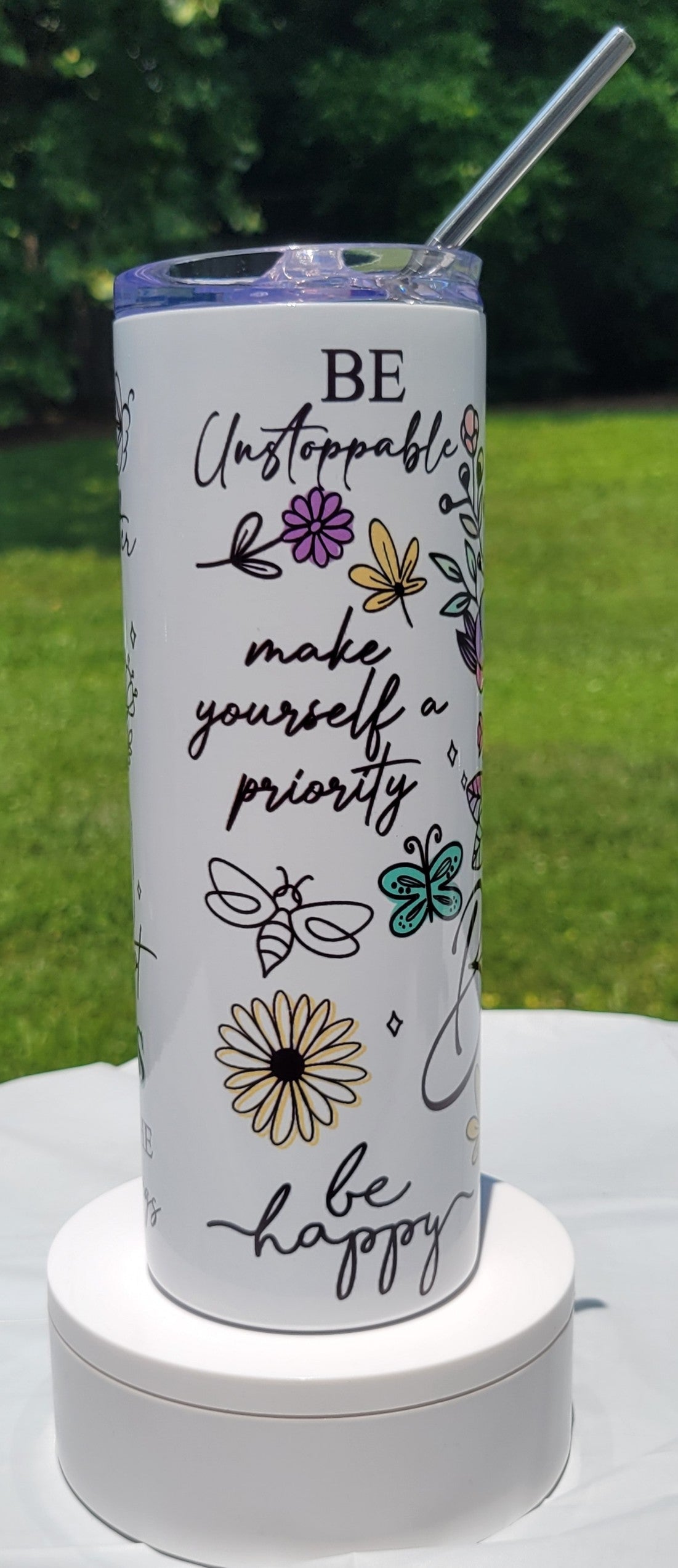 Tumbler with motivational quote and floral designs on a white surface with greenery in the background